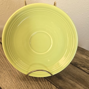 May include: A round, light yellow ceramic saucer featuring a series of concentric circular ridges embossed along the rim. The saucer has a smooth, glossy finish and a central indentation designed to hold a matching cup securely in place.