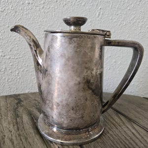 May include: A tarnished silver-toned metal teapot featuring a cylindrical body, a curved spout, a hinged lid with a small knob handle, and a sturdy side handle. The surface shows visible signs of oxidation and wear, creating a dark, mottled patina.