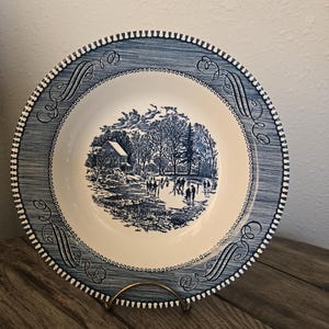 Può includere: Un piatto rotondo in ceramica con un motivo blu e bianco. Il centro raffigura un paesaggio invernale con una casa, alberi e figure sul ghiaccio. Il bordo presenta una cornice blu testurizzata con decorazioni a volute e una linea tratteggiata.