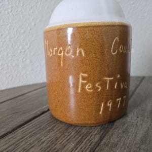 Puede incluir: Un tarro de cerámica vintage con cuerpo marrón y tapa blanca. El tarro tiene las palabras "Morgan County Festival 1977" grabadas en el lateral. El tarro está sobre una superficie de madera.