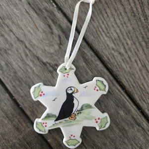 May include: A white ceramic star-shaped ornament featuring a hand-painted puffin standing on a green grassy landscape. The design includes holly leaves with red berries at each point of the star and a white ribbon loop for hanging the decoration.