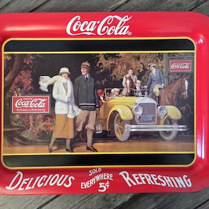May include: A vintage Coca-Cola serving tray with a red border and a central image depicting people in vintage attire near a yellow car and a horse. The tray has the Coca-Cola logo and the words "Delicious" and "Refreshing".