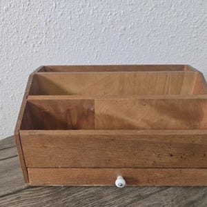 May include: A light brown wooden desk organizer featuring two tiered open compartments for document storage and a single bottom drawer equipped with a small, round, white ceramic knob for easy access to stored items and supplies.