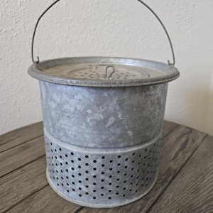 May include: A galvanized metal bucket with a perforated lower section and a lid. The bucket has a wire handle and is sitting on a wooden surface. The bucket is likely used for carrying or storing items.