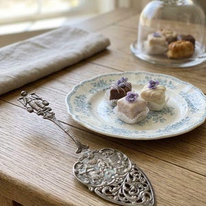 May include: An ornate silver-toned metal cake server with an embossed handle and a wide, decorative triangular blade featuring intricate relief patterns, positioned beside a white ceramic plate with blue floral details holding three small square cakes.