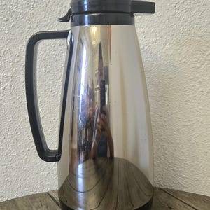 May include: A tall, cylindrical thermal carafe featuring a polished, reflective stainless steel body. It includes a black plastic handle, a black lid with a pouring spout, and a black base. The surface shows a clear reflection of the surroundings.