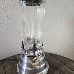 May include: A vintage-style blender featuring a tall, clear, vertically ribbed glass pitcher. The base is constructed from polished chrome with a tiered, circular design. A black plastic lid sits on top, and black accents secure the glass pitcher.