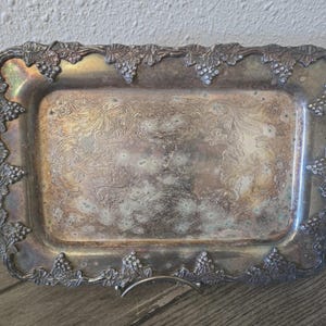 May include: A rectangular silver-toned metal tray featuring an ornate border decorated with embossed grape clusters and vine leaves. The center of the tray displays a detailed, etched floral scrollwork pattern with visible tarnishing.