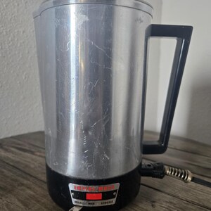 Könnte beinhalten: Ein Vintage-Elektro-Kaffeezubereiter mit silberfarbenem Gehäuse, schwarzem Sockel und Griff. Der Perkolator hat einen Glasdeckel und ein Netzkabel mit einem spiralförmigen Abschnitt. Das Bedienfeld zeigt "Mild, Med, Strong."
