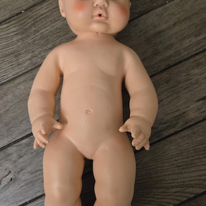 May include: A baby doll with a smooth, light-toned vinyl body, molded hair, and painted facial features. The doll has large, wide-set eyes, arched eyebrows, and rosy pink cheeks. It features a small, open mouth and a defined belly button.