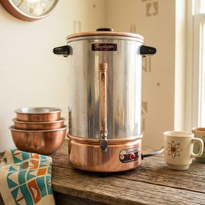 Puede incluir: Una cafetera percoladora Tricolator vintage con cuerpo cilíndrico plateado pulido, base de color cobre y asas laterales negras. La unidad cuenta con un visor de nivel vertical, un grifo frontal y una pequeña luz indicadora roja.