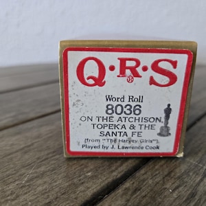 May include: A vintage QRS Word Roll box, featuring a red and white label with the text "QRS" and "8036 ON THE ATCHISON, TOPEKA & THE SANTA FE." The box is made of cardboard and sits on a wooden surface.