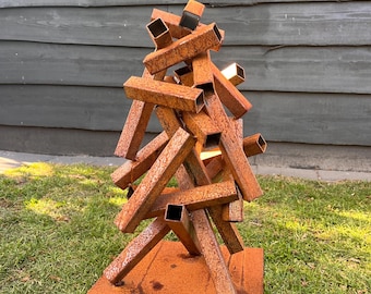 May include: A weathered metal sculpture made from stacked, square tubes. The sculpture rests on a square metal base, positioned on a grassy lawn. A yellow butterfly decoration is fixed to the wooden wall behind it.