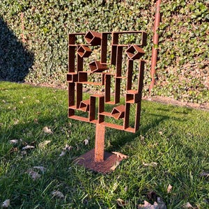 Op de afbeelding: Een roestbruine metalen sculptuur met een geometrisch ontwerp. De sculptuur bestaat uit rechthoekige en vierkante vormen, geplaatst op een vierkante basis. Het staat op een grasveld voor een groene haag.