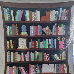 Peut inclure: Une courtepointe représentant une bibliothèque remplie de livres en tissu colorés. La bibliothèque a cinq étagères et est faite de tissu brun. Il y a un vase avec des fleurs sur l'étagère supérieure et un cadre photo sur la troisième étagère.