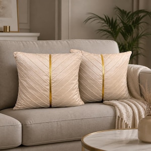 Peut inclure: Deux coussins carrés beiges avec une bande verticale dorée et un motif cousu en diagonale sur un canapé gris. Une couverture crème et un petit vase avec des fleurs séchées sont également visibles.