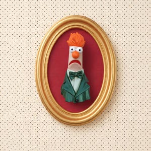 May include: A three-dimensional bust of the Muppet character Beaker, featuring bright orange hair, a pale face, and a wide mouth, wearing a dark green suit jacket and a matching bow tie, encased within an oval gold-toned decorative frame.
