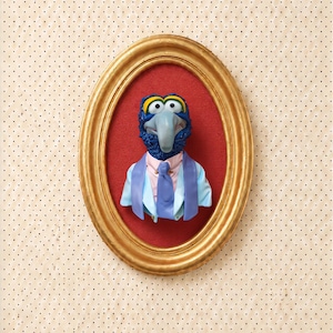 May include: A three-dimensional bust of the character Gonzo from The Muppets, featuring a blue face, large nose, and yellow eyelids. The figure wears a light blue blazer, pink shirt, and blue tie, displayed inside an oval gold frame on red.