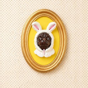 May include: A three-dimensional wall art piece featuring a dark brown pug head wearing a white bunny ear hood with pink inner ears. The pug is centered within an oval gold frame against a bright yellow background, creating a whimsical portrait.