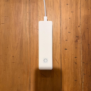 May include: A rectangular white electronic device with a smooth matte finish and a single circular button on the front surface. A white power cable extends from the top edge of the unit, which is positioned vertically against a wooden background.