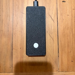May include: A rectangular black electronic device with a textured surface and a single circular white button near the bottom. A black braided cable is connected to the top center of the unit, extending upwards from the main body of the device.