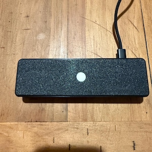 May include: A rectangular black electronic device with a textured, glittery surface and a single circular white button in the center. A black braided power cable is plugged into the top right side of the unit, extending upwards from the device.