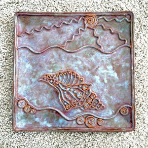May include: Square ceramic dish with a raised design featuring a bird in flight, sun, and wavy lines. The dish has a mottled blue and brown glaze with copper-colored accents. The edges are raised and the overall design is artistic.