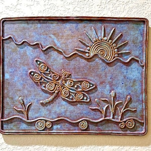 May include: A rectangular wall plaque featuring a relief design of a dragonfly in flight above water with reeds and a sun. The piece has a mottled blue and copper-toned finish with raised spiral patterns forming the insect, sun, and landscape elements.