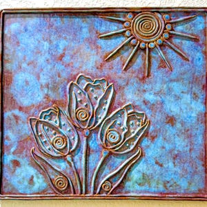 May include: A square metal wall plaque featuring a raised relief design of three tulips and a sun. The surface has a mottled blue and copper-toned patina. The sun is depicted with a spiral center and radiating rays, while the tulips feature spiral details.
