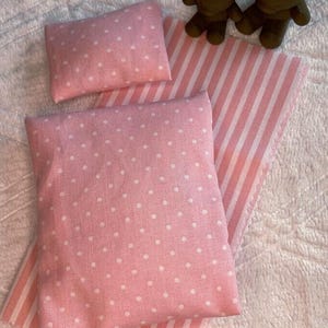 Könnte beinhalten: Ein Miniatur-Bettwäscheset bestehend aus einer rosa-weiß gestreiften Matratzenauflage, einem rosa Kissen mit weißen Punkten und einer größeren rosa Decke mit weißen Punkten. Zwei kleine braune Bärenfiguren stehen oben am Set.