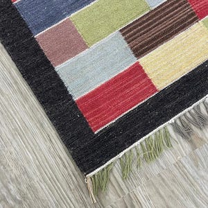 Könnte beinhalten: Ein farbenfroher rechteckiger Teppich mit einem Patchwork-Design. Der Teppich hat einen schwarzen Rand und eine Vielzahl rechteckiger Blöcke in Rot-, Blau-, Grün-, Braun- und Gelbtönen. Der Teppich hat einen Fransen aus grünen Fäden.