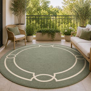 Könnte beinhalten: Ein runder Outdoor-Teppich in gedämpftem Grün mit einem cremefarbenen geometrischen Rand. Der Teppich liegt auf einer gefliesten Terrasse, mit Gartenmöbeln und Pflanzen im Hintergrund. Das Design des Teppichs verleiht dem Außenbereich Eleganz.