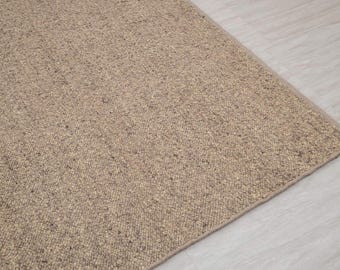 May include: A corner view of a beige woven rug. The rug has a textured surface with small flecks of darker color. The rug is placed on a light-colored wooden floor.