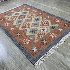 Könnte beinhalten: Rechteckiger Teppich mit geometrischem Muster in Orange, Blau, Gelb und Rot. Er hat einen blauen Rand und weiße Fransen. Das zentrale Design zeigt Rauten. Abmessungen in Zentimetern.