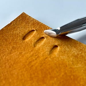 May include: Close-up of a leather crafting tool in use. The tool is silver and is pressing into a piece of tan leather, creating a series of evenly spaced impressions. The leather has a textured surface.