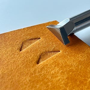 May include: Close-up of a leather crafting tool with a textured, angled head. The tool is pressed into a piece of tan leather, creating a detailed, angled pattern. The leather has a natural, textured appearance.