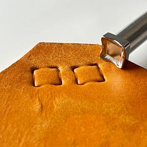 以下が含まれることがあります： オレンジブラウンの革に正方形の刻印を作る革細工ツールのクローズアップ。ツールは正方形のヘッドとシルバーのハンドルが付いています。革は質感のある表面です。