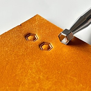 May include: Close-up of leather crafting tools. A hexagonal stamp is pressed into a piece of textured, orange-brown leather, creating a geometric design. The tool is silver, and the leather has a natural, aged appearance. The image highlights the process of leatherwork.