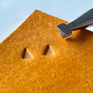 Pode incluir: Close-up de um projeto de artesanato em couro. Uma ferramenta de metal é usada para estampar um padrão triangular texturizado em um pedaço de couro bronzeado. Dois triângulos estampados são visíveis, mostrando os detalhes do trabalho.