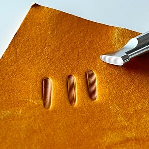 Puede incluir: Primer plano de un trozo de cuero curtido con tres impresiones verticales texturizadas. Una herramienta de metal plateado con un borde biselado se coloca cerca del cuero, lo que sugiere un proceso de trabajo del cuero. El cuero tiene un tono marrón dorado cálido.