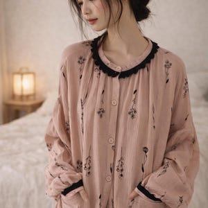 May include: A light pink pajama set with a floral pattern. The long-sleeved top has a black lace trim around the collar and cuffs, and black trim on the pockets. The set is button-down and appears to be made of a soft, lightweight fabric.