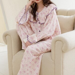 May include: A light pink floral pajama set. The long-sleeved top has a ruffled collar and cuffs trimmed with purple. The matching pants are wide-legged. The pajamas are worn while seated on a cream-colored armchair.