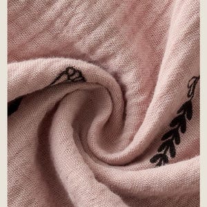 May include: Close-up of a dusty rose fabric with a woven texture. The fabric has a delicate, slightly wrinkled appearance. Black leaf-like designs are printed along the edge. The word "FABRIC" is printed in a serif font at the top.