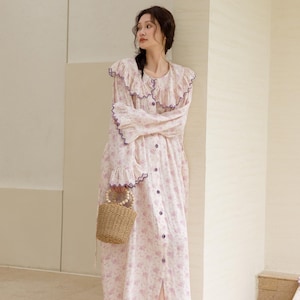 May include: A light pink floral print dress with a ruffled collar and sleeves, accented with purple trim. The dress has a button-down front and is paired with a small woven handbag. The model is wearing brown sandals.
