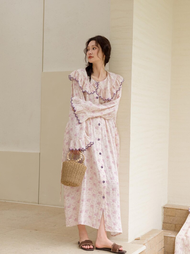 May include: A light pink floral print dress with a ruffled collar and sleeves, accented with purple trim. The dress has a button-down front and is paired with a small woven handbag. The model is wearing brown sandals.