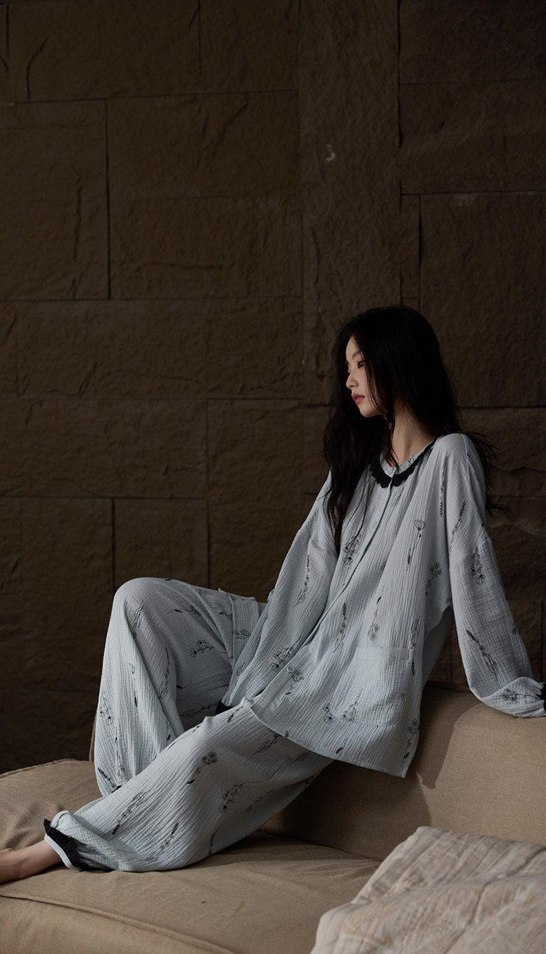 May include: Light blue pajama set with a floral pattern. The long-sleeved top has a black trim around the collar and cuffs. The matching pants are loose-fitting. The fabric appears to be a soft, textured material.