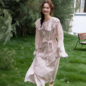 May include: A light pink floral print nightgown with a ruffled collar and cuffs. The long-sleeved gown features a button-down front, a waist tie, and purple trim. The model is wearing brown sandals.