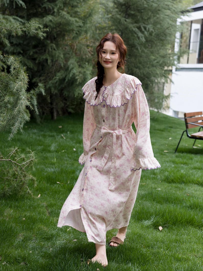 May include: A light pink floral print nightgown with a ruffled collar and cuffs. The long-sleeved gown features a button-down front, a waist tie, and purple trim. The model is wearing brown sandals.