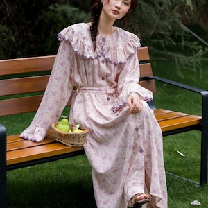 May include: A light pink floral print dress with long sleeves, a ruffled collar, and a matching belt. The dress has purple trim and buttons down the front. A small woven basket with fruit sits nearby. The person is wearing brown sandals.