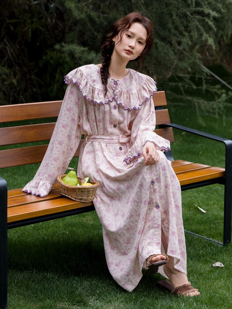 May include: A light pink floral print dress with long sleeves, a ruffled collar, and a matching belt. The dress has purple trim and buttons down the front. A small woven basket with fruit sits nearby. The person is wearing brown sandals.